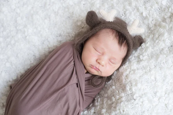 A cute little baby in a knitted hat with deer horns and a brown blanket sleeps on a white boucl bedspread at home. Health and motherhood