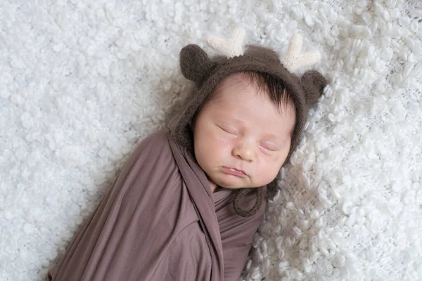 A cute little baby in a knitted hat with deer horns and a brown blanket sleeps on a white boucl bedspread at home. Health and motherhood