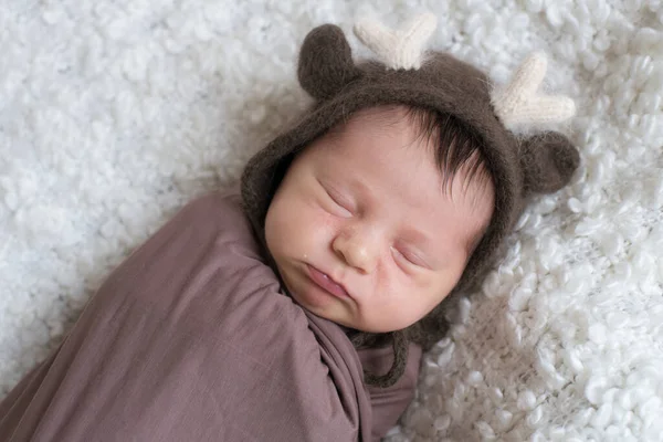 A cute little baby in a knitted hat with deer horns and a brown blanket sleeps on a white boucl bedspread at home. Health and motherhood