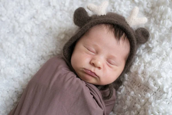 A cute little baby in a knitted hat with deer horns and a brown blanket sleeps on a white boucl bedspread at home. Health and motherhood