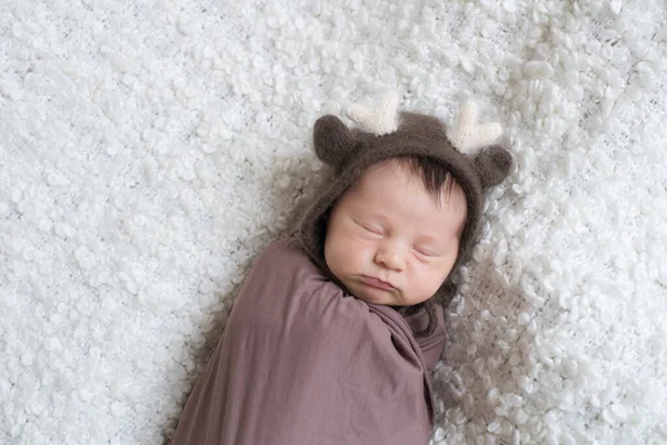 A cute little baby in a knitted hat with deer horns and a brown blanket sleeps on a white boucl bedspread at home. Health and motherhood