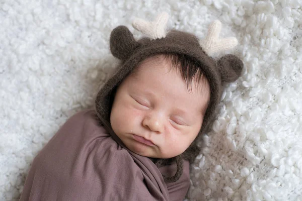 A cute little baby in a knitted hat with deer horns and a brown blanket sleeps on a white boucl bedspread at home. Health and motherhood