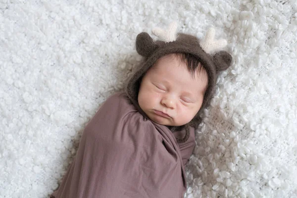 A cute little baby in a knitted hat with deer horns and a brown blanket sleeps on a white boucl bedspread at home. Health and motherhood