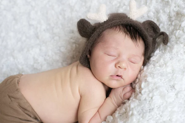 A cute little baby in a knitted hat with deer horns sleeps on a white boucl bedspread at home. Health and motherhood