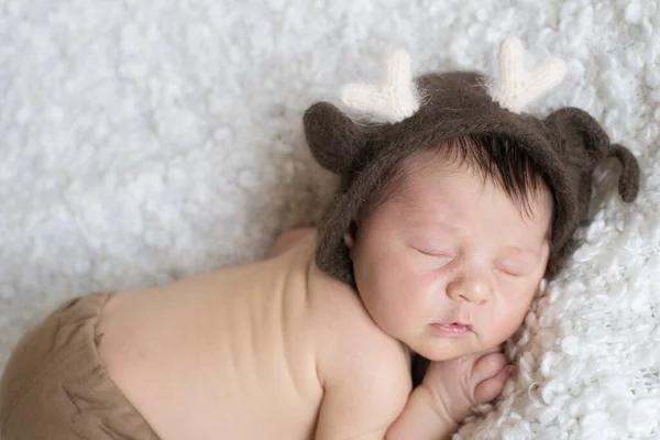 A cute little baby in a knitted hat with deer horns sleeps on a white boucl bedspread at home. Health and motherhood