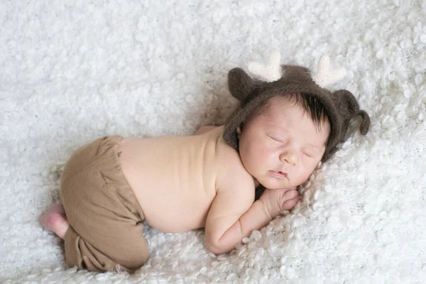 A cute little baby in a knitted hat with deer horns sleeps on a white boucl bedspread at home. Health and motherhood