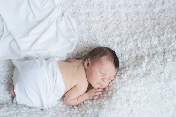 A cute little baby in a white blanket sleeps on a white boucl bedspread at home. Health and motherhood