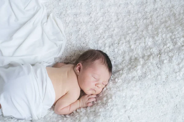 A cute little baby in a white blanket sleeps on a white boucl bedspread at home. Health and motherhood