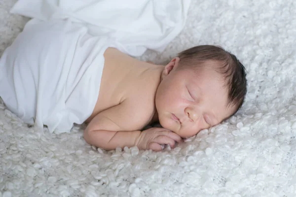 A cute little baby in a white blanket sleeps on a white boucl bedspread at home. Health and motherhood