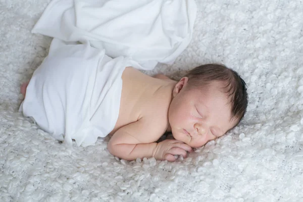 A cute little baby in a white blanket sleeps on a white boucl bedspread at home. Health and motherhood