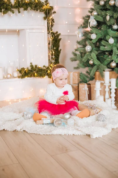 A little girl under one year old in an airy dress in a room decorated for Christmas,  near the Christmas tree among the pillows, gifts, garlands and pine needles. Christmas mood.