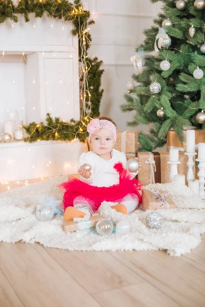 A little girl under one year old in an airy dress in a room decorated for Christmas,  near the Christmas tree among the pillows, gifts, garlands and pine needles. Christmas mood.