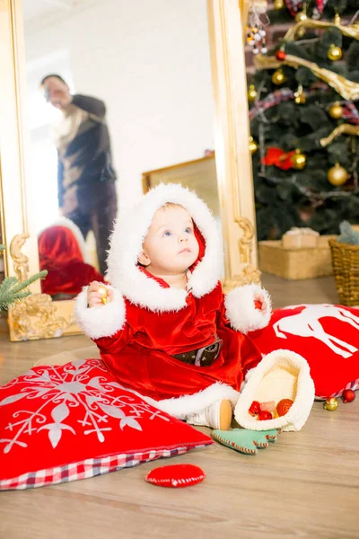 一个一岁以下的小女孩穿着圣诞老人的衣服 住在圣诞树旁的一间装饰过圣诞节的房间里 身边有枕头 花环和松针 圣诞气氛 儿童与圣诞节 — 图库照片