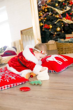 Noel için süslenmiş bir odada Noel Baba kılığındaki küçük bir kız çocuğu yastıkların, hediyelerin çelenklerin ve çam iğnelerinin arasında Noel ağacının yanında. Noel havası. Çocuklar ve Noel