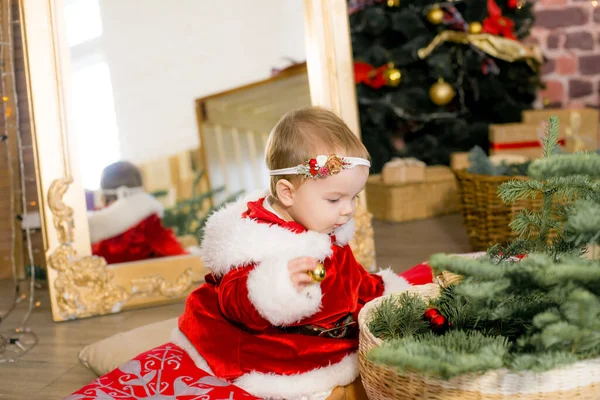 A little girl under one year old dressed as Santa Claus in a room decorated for Christmas, near the Christmas tree among the pillows, gifts, garlands and pine needles. Christmas mood. Children and Christmas