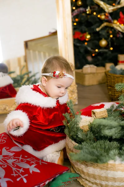 1歳未満の女の子は クリスマスのために装飾された部屋でサンタクロースの服を着て 贈り物 ガーランド 松葉の間のクリスマスツリーの近くにあります クリスマス気分 子供とクリスマス — ストック写真