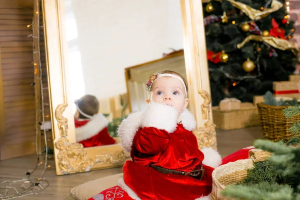 1歳未満の女の子は クリスマスのために装飾された部屋でサンタクロースの服を着て 贈り物 ガーランド 松葉の間のクリスマスツリーの近くにあります クリスマス気分 子供とクリスマス — ストック写真