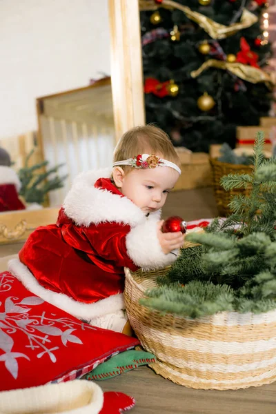 一个一岁以下的小女孩穿着圣诞老人的衣服 住在圣诞树旁的一间装饰过圣诞节的房间里 身边有枕头 花环和松针 圣诞气氛 儿童与圣诞节 — 图库照片