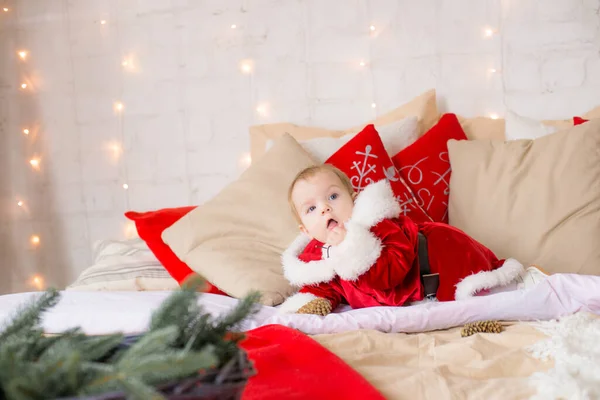 A little girl under one year old dressed as Santa Claus on a large bed in a room decorated for Christmas, among the pillows of garlands and pine needles. Christmas mood. Children and Christmas