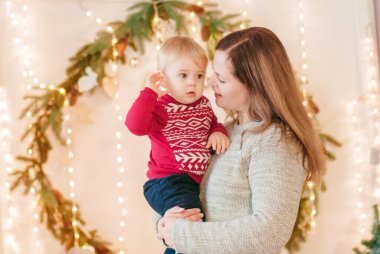 Genç, sarı saçlı bir anne ve küçük oğlu Noel için çam iğneleri ve parlak çelenklerle süslenmiş bir odada kollarında. Noel havası