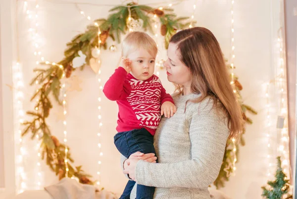 A young blond-haired mother with her little son in her arms in a room decorated with pine needles and sparkling garlands for Christmas. Christmas mood