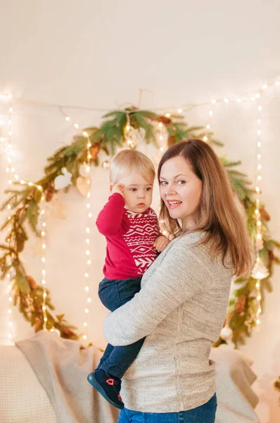 A young blond-haired mother with her little son in her arms in a room decorated with pine needles and sparkling garlands for Christmas. Christmas mood