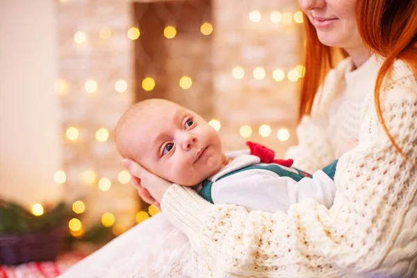 A young red-haired mother with her little baby in her arms in a room decorated with pine needles and sparkling garlands for Christmas. Christmas mood