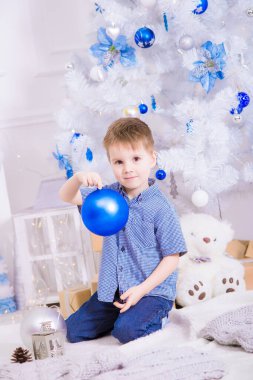 Noel için dekore edilmiş bir odada, beyaz bir Noel ağacının yanında mavi gömlekli yakışıklı bir çocuk. Aile Noel 'i Noel havası