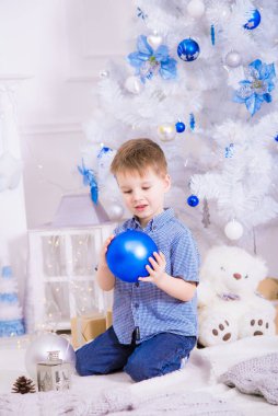 Noel için dekore edilmiş bir odada, beyaz bir Noel ağacının yanında mavi gömlekli yakışıklı bir çocuk. Aile Noel 'i Noel havası