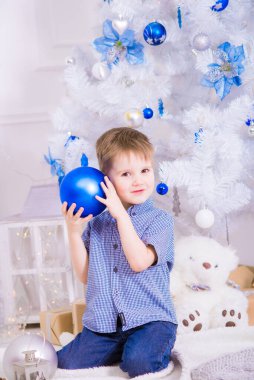 Noel için dekore edilmiş bir odada, beyaz bir Noel ağacının yanında mavi gömlekli yakışıklı bir çocuk. Aile Noel 'i Noel havası