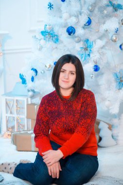Noel için dekore edilmiş bir odada, beyaz bir Noel ağacının yanında kırmızı kazaklı, koyu renk saçlı genç bir kadın. Aile Noel 'i Noel havası