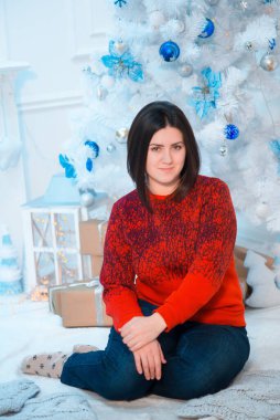 Noel için dekore edilmiş bir odada, beyaz bir Noel ağacının yanında kırmızı kazaklı, koyu renk saçlı genç bir kadın. Aile Noel 'i Noel havası