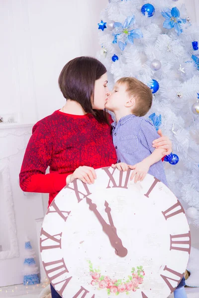 白いクリスマスツリーとクリスマスのために装飾された部屋で大きな時計の近くに彼女の息子と暗い髪を持つ若い母親 クリスマスだ クリスマス気分 — ストック写真