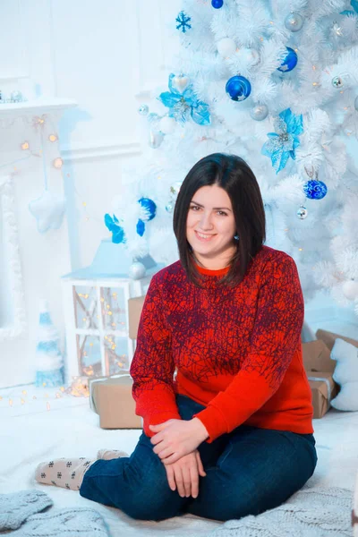 Noel için dekore edilmiş bir odada, beyaz bir Noel ağacının yanında kırmızı kazaklı, koyu renk saçlı genç bir kadın. Aile Noel 'i Noel havası