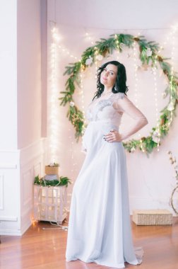 Noel için çam iğneleri ve ışıl ışıl çelenklerle süslenmiş bir odada koyu renk saçlı hamile bir kadın. Noel havası. Hamilelik