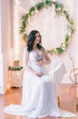 Noel için çam iğneleri ve ışıl ışıl çelenklerle süslenmiş bir odada koyu renk saçlı hamile bir kadın. Noel havası. Hamilelik