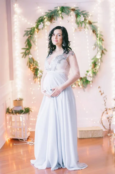 Young pregnant woman with dark hair in airy dress in a room decorated with pine needles and sparkling garlands for Christmas. Christmas mood . Pregnancy