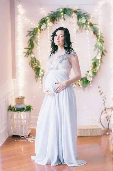 Young pregnant woman with dark hair in airy dress in a room decorated with pine needles and sparkling garlands for Christmas. Christmas mood . Pregnancy