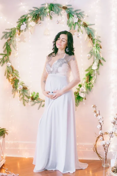 松の針とクリスマスのための輝くガーランドで飾られた部屋で 風通しの良いドレスで暗い髪を持つ若い妊婦 クリスマス気分 — ストック写真