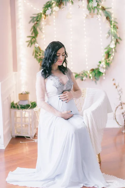 Young pregnant woman with dark hair in airy dress in a room decorated with pine needles and sparkling garlands for Christmas. Christmas mood . Pregnancy