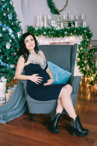 ろうそく 松葉やガーランドやクリスマスツリーで飾られた暖炉の近くのドレスで暗い髪を持つ若い妊婦 クリスマス気分 — ストック写真