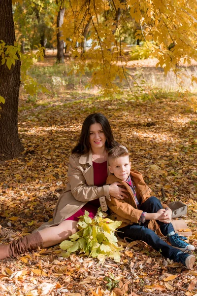 一位年轻的母亲 一头乌黑的头发 穿着米黄色的秋天雨衣 和她的儿子在一个阳光灿烂的秋天公园里 在一片黄色的树叶中 秋天在公园里散步 秋天的心情母性 在一起的时间 — 图库照片