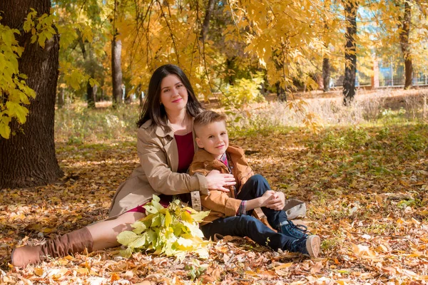 黄色の葉の間の日当たりの良い秋の公園で彼女の息子とベージュの秋のレインコートで暗い髪の若い母親 公園の秋の散歩 秋の気分 一緒に時間を — ストック写真