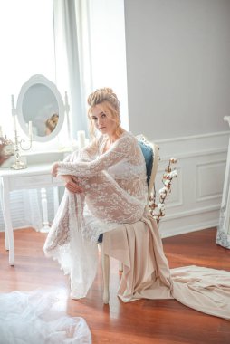 Sabah düğün saç stili ve makyajlı genç ve güzel bir gelin yatak odası masasının yanında, yere kadar uzanan beyaz dantelli bir ceketle. düğün sabahı