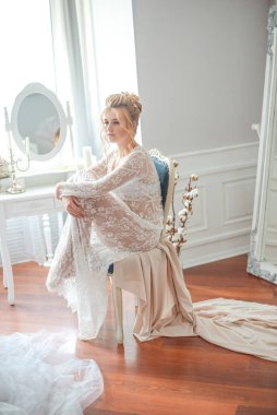 Sabah düğün saç stili ve makyajlı genç ve güzel bir gelin yatak odası masasının yanında, yere kadar uzanan beyaz dantelli bir ceketle. düğün sabahı