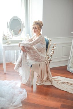 Sabah düğün saç stili ve makyajlı genç ve güzel bir gelin yatak odası masasının yanında, yere kadar uzanan beyaz dantelli bir ceketle. düğün sabahı
