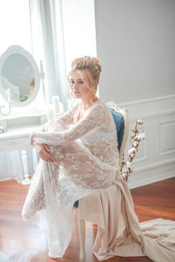 Sabah düğün saç stili ve makyajlı genç ve güzel bir gelin yatak odası masasının yanında, yere kadar uzanan beyaz dantelli bir ceketle. düğün sabahı