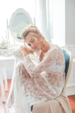 Sabah düğün saç stili ve makyajlı genç ve güzel bir gelin yatak odası masasının yanında, yere kadar uzanan beyaz dantelli bir ceketle. düğün sabahı