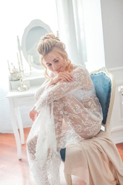 Sabah düğün saç stili ve makyajlı genç ve güzel bir gelin yatak odası masasının yanında, yere kadar uzanan beyaz dantelli bir ceketle. düğün sabahı