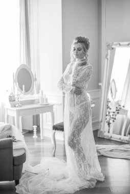 Sabah düğün saç stili ve makyajlı genç ve güzel bir gelin yatak odası masasının yanında, yere kadar uzanan beyaz dantelli bir ceketle. düğün sabahı